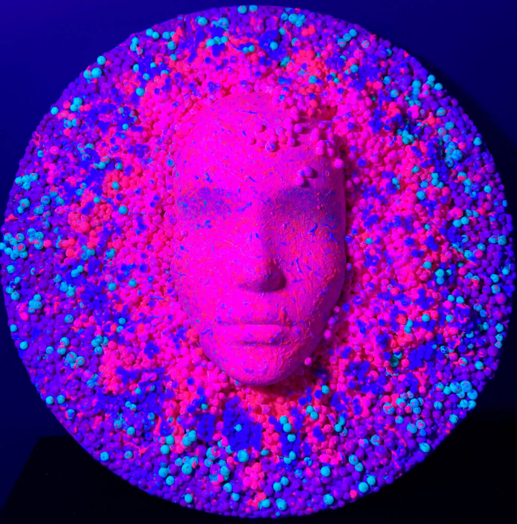 High-relief masks depicting the face losing it shape, rendered in a palette of purple and violet, with a pointillism effect created by small spheres forming the artwork